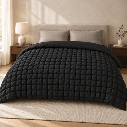 Schwarze Bettdecke mit kleinen, bauschigen Steppquadraten, schräg von vorne in einem hellen Schlafzimmer fotografiert.